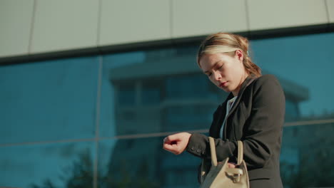 side view of white lady on way to lecture checking handbag to retrieve keys, standing near modern glass building reflecting her image, sunlight illuminating her face and professional attire