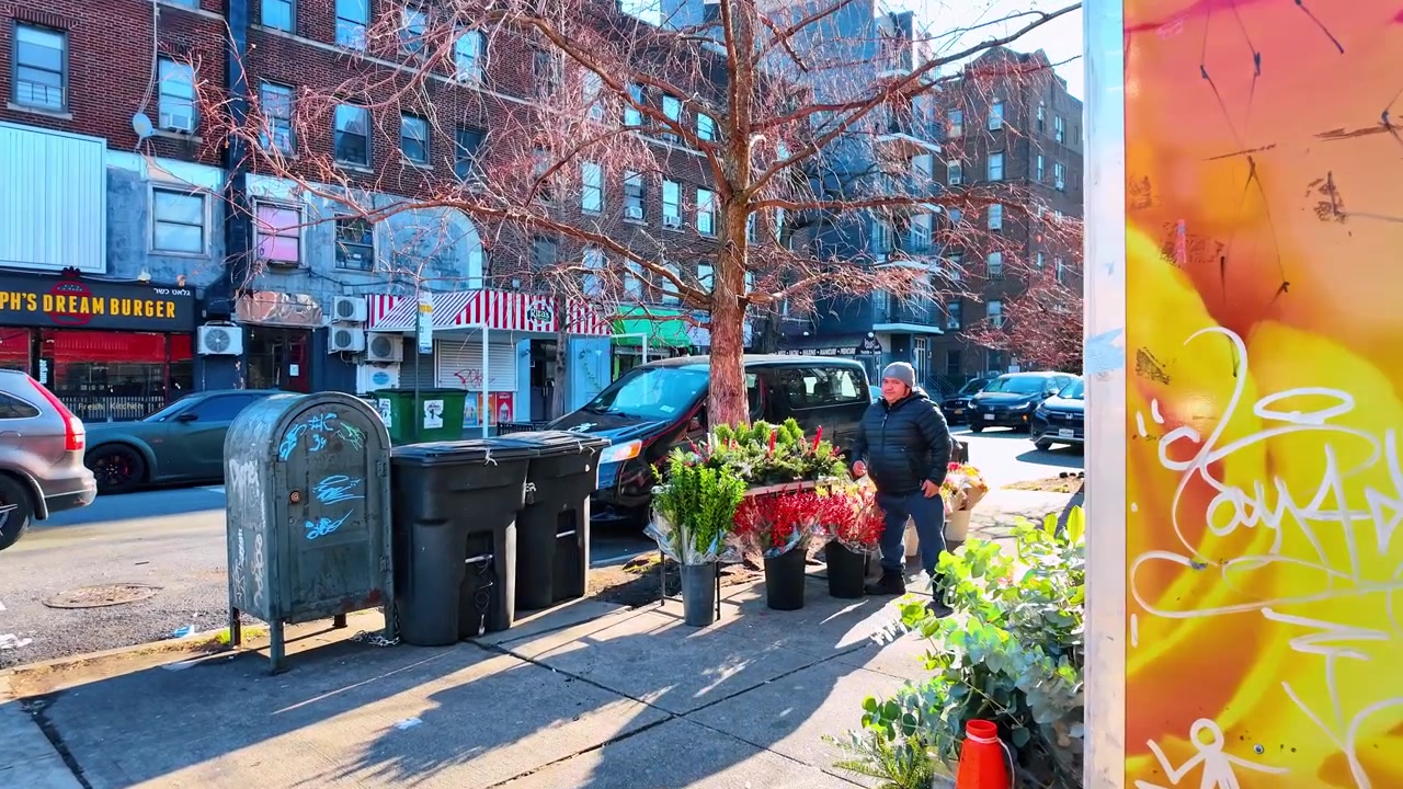 New York, USA, 20 december 2025: Gatuförsäljare arrangerar blommor bredvid en graffitivägg. En man står bland hinkar med snittgrönt på en solig stadstrottoar.