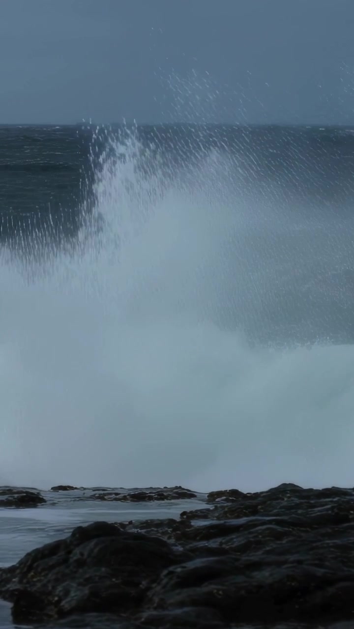Vertical video: Approaching swell, large breaking wave crashing rocky shoreline, spraying foam