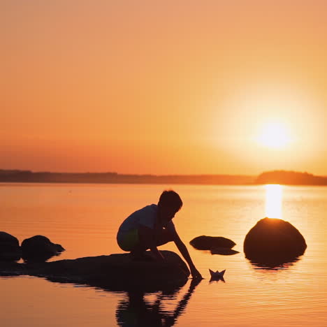 child with paper ship at sunset. little boy sitting on a stone in the lake and putting origami boat on the water. orange background of the evening sky. slow motion.