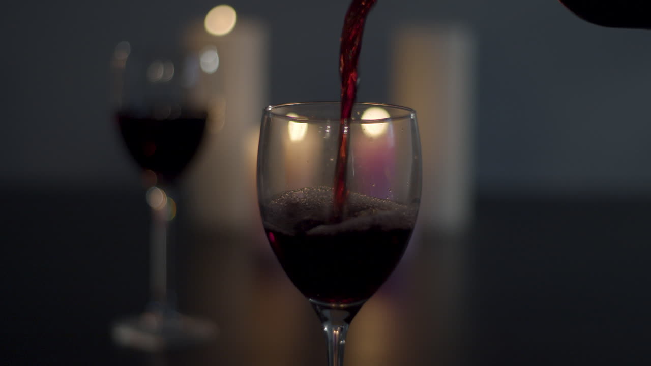 Vino rosso versato in un bicchiere con un bicchiere pieno e candele accese sullo sfondo