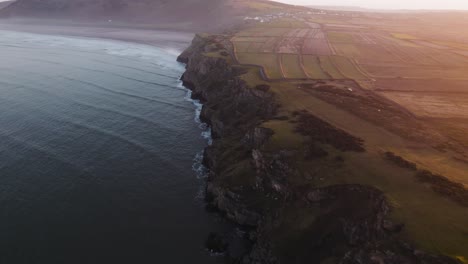 aerial: dolly along cliff face coastline during hazy sunrise, rhossili gower, 4k drone