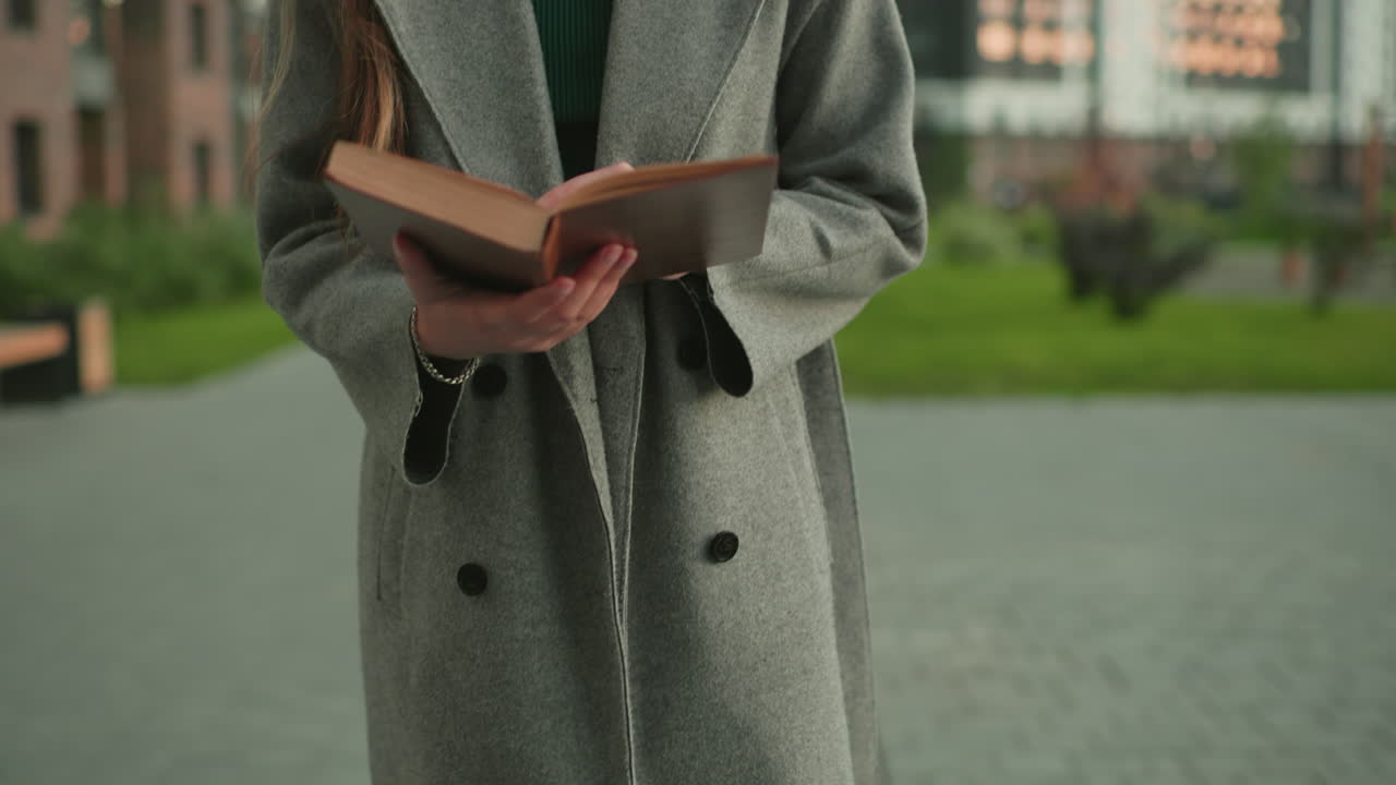 Close up of lady in gray coat holding book while moving backward outdoors with blur urban background, showing focus, calm mood, lifestyle, and learning activity