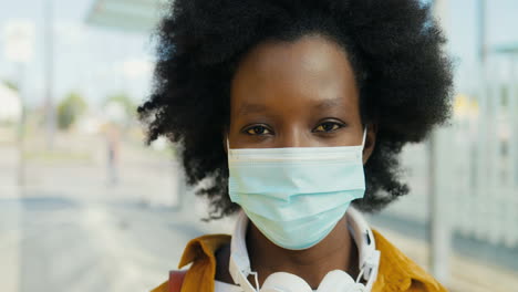 retrato de una joven viajera afroamericana con máscara médica y auriculares mirando la cámara al aire libre