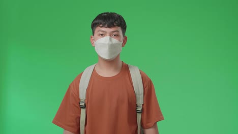 the front view of asian boy student wearing a mask, backpack and brown t-shirt walking to school on green screen chroma key.