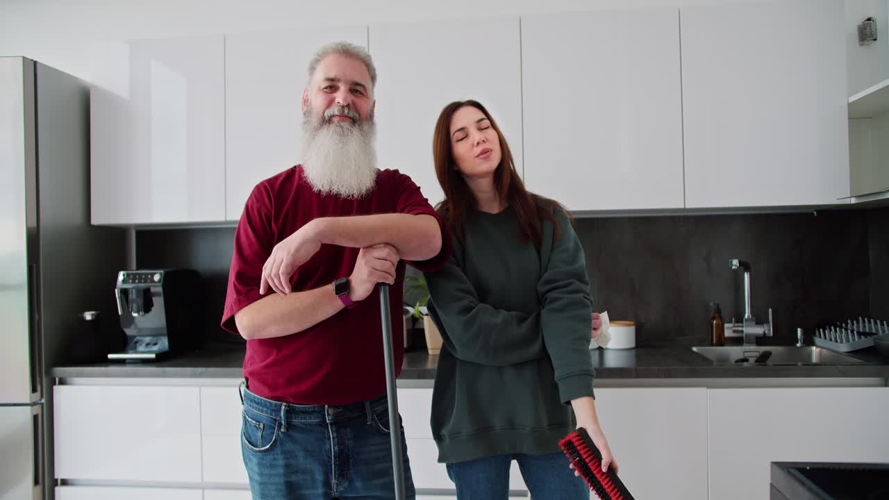 Portrait of a happy elderly man with gray hair and a lush beard in a red T-shirt who poses with his adult brunette daughter in a green sweater while cleaning in a modern kitchen