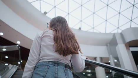 back view of young lady on moving escalator in modern mall, phone in back pocket, hand on rail, with other people ascending in front, showcasing everyday shopping experience and urban lifestyle