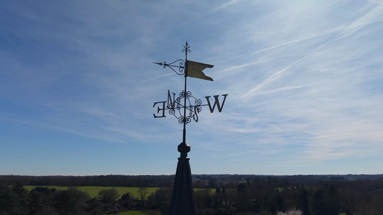 Church weather vane drone orbit at St Mary's, Hertingfordbury, England. Near-360 aerial rotation around spire finial as sunlight catches the gilded metal against blue sky