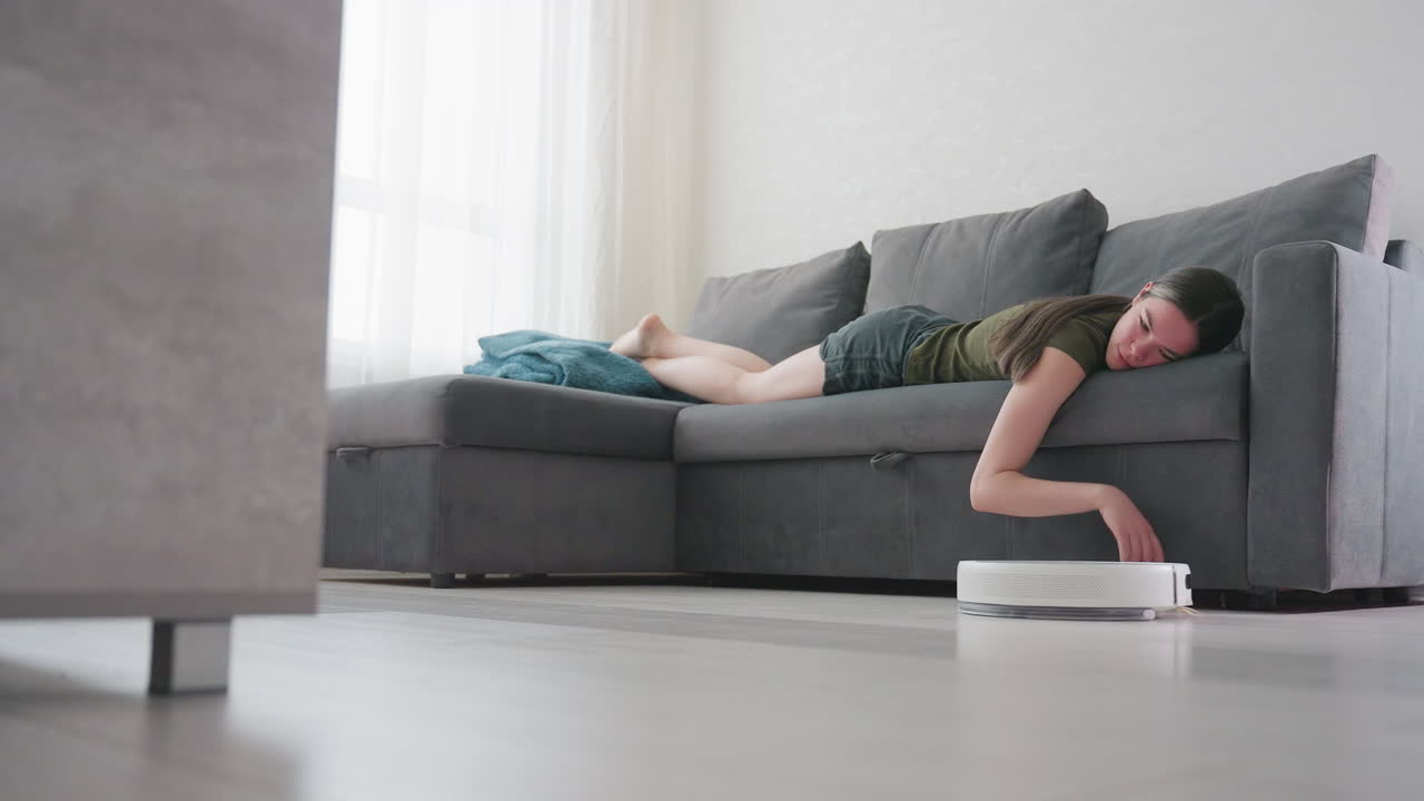 Woman lies on gray couch with relaxed posture, hand resting on floor as robot vacuum cleaner passes by, illustrating smart home cleaning, effortless automation, and modern lifestyle comfort indoors