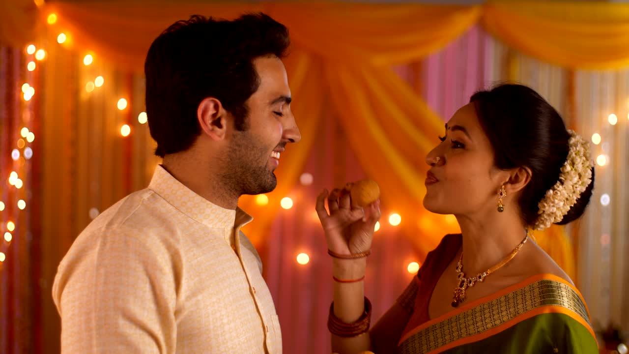 Husband Wife feeding sweets to each other on the festive occasion - Colorful background with lights