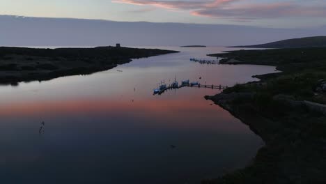 Aerial-drone-shot-dropping-behind-hills-as-the-reflection-of-fire-sunset-sets-in-the-distance-across-the-water