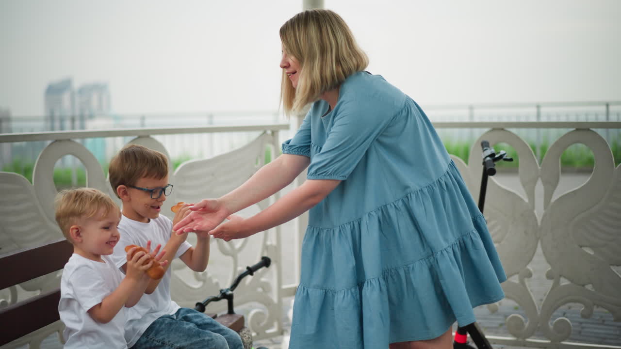 una mujer con una túnica azul ofrece un rollo de salchichas a sus dos hijos. el niño mayor toma un bocado mientras el más joven mira con curiosidad, un scooter negro está estacionado cerca