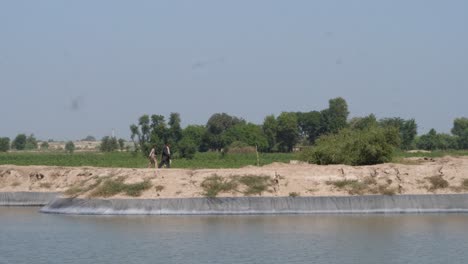 shot of a center pivot irrigation sprinkler system on other side of a river over farmlands with locals passing by in punjab