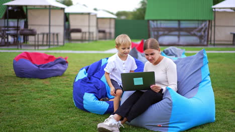 mother and little son looking at laptop sitting in bean bag chair. chatting and laughing spending time outdoors in summer.