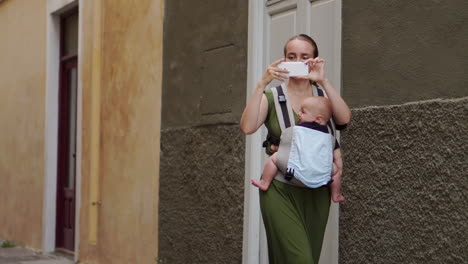 A-young-mother-with-her-baby-in-a-kangaroo-backpack-captures-their-journey-through-photos-on-a-mobile-phone.-As-they-walk,-she-glances-at-the-phone-screen