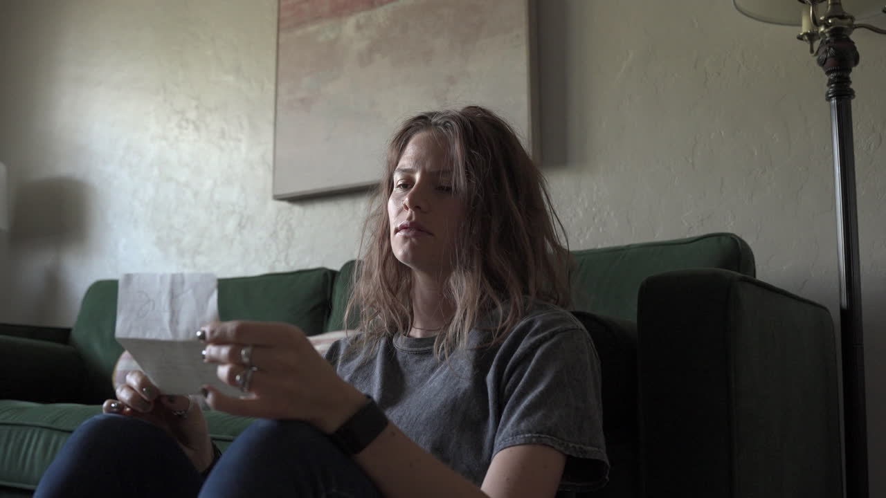 A woman sits on the floor in front of a green couch, reading a love letter aloud. Soft backlighting highlights her figure as the camera slowly turns around her, capturing the moment's warmth.