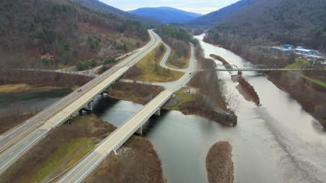 Volando-Sobre-El-Valle-De-Un-Río-Con-Dos-Puentes-Que-Cruzan-El-Agua,-Un-Pequeño-Pueblo-Y-Montañas-A-Fines-Del-Otoño