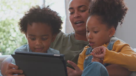 Madre-Con-Hijos-Jugando-O-Transmitiendo-A-Una-Tableta-Digital-Sentados-Juntos-En-Un-Sofá-En-Casa