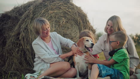 woman and her daughter with children play with leashed dog outdoors, smiling joyfully near hay bale in open field, casual family gathering during golden hour