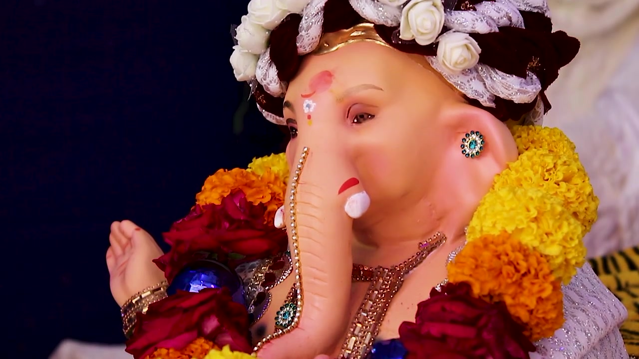 Small Ganesha idol side profile closeup wearing white rose turban, marigold garland dark background Ganesh Chaturthi festival India.