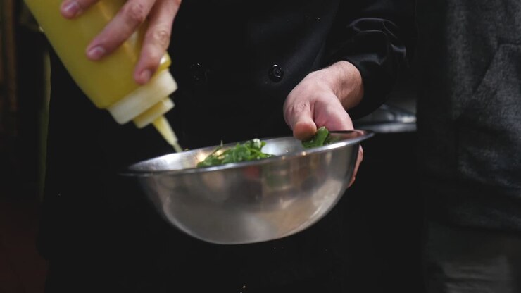 Chef professionista prepara un'insalata nella cucina di un ristorante, aggiungendo il condimento all'insalata in una ciotola di miscelazione in acciaio inossidabile, angolazione fissa semi-ravvicinata, al rallentatore.