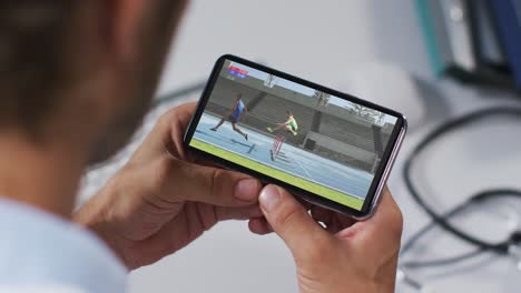 compuesto de un médico masculino viendo un evento de atletismo en un teléfono inteligente