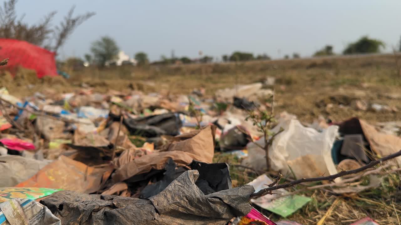 Close-up of scattered plastic bags and mixed waste along a roadside verge, with dry grass in the foreground and distant vehicles softly blurred across an open landscape