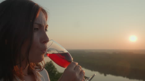 side view of woman admiring red wine in glass as she gently sips while watching sunset over river valley, soft golden light illuminates her face, insects fly around