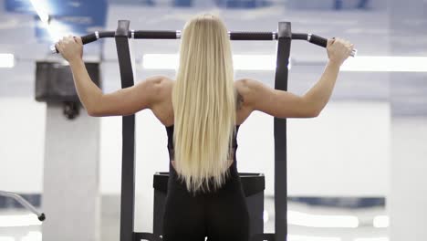 Strong-female-bodybuilder-with-blond-hair-with-muscular-arms-performing-pulls-up-on-a-horizontal-bar-at-the-gym.-Backside-footage