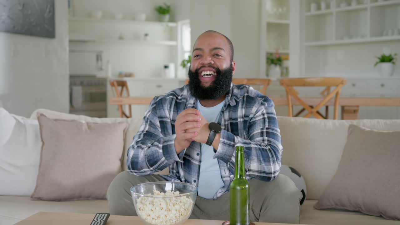Voksen afrikansk mand iført flannel ser tv derhjemme, reagerer, klapper, rækker ud efter øl og popcorn