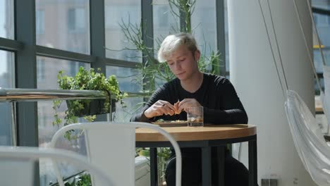 young man with blond hair enjoying a sandwich and iced coffee at a cafe table by a window with city views, natural light, and potted plants. relaxed and casual dining scene