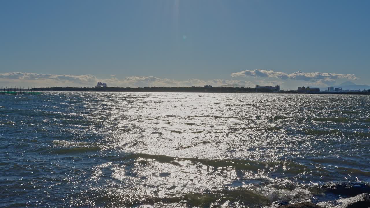 A shimmering path of sunlight on choppy water with a low-lying distant city skyline and faint clouds on the horizon