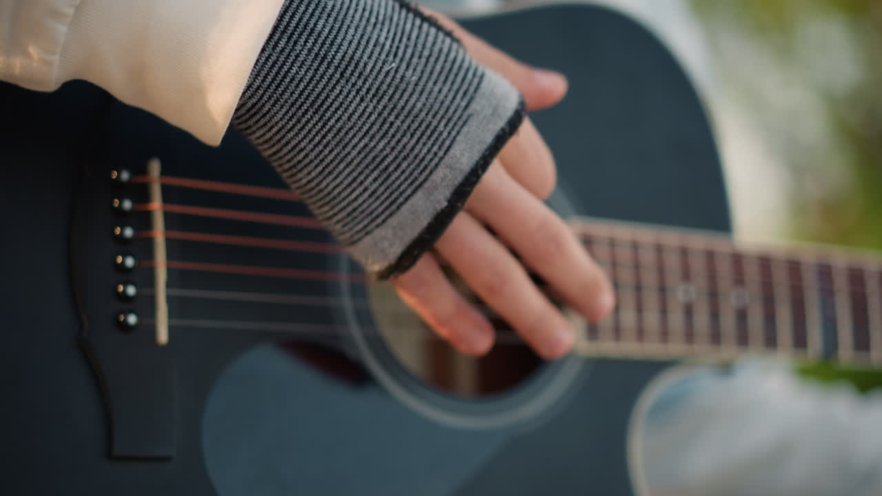 primo piano di mani su chitarra acustica, guanto senza dita e dita vicino alle corde, cantautore indie che si esercita con un delicato strumming su una panchina del parco durante l'ora d'oro, messa a fuoco ridotta, luce naturale calda, rilassato
