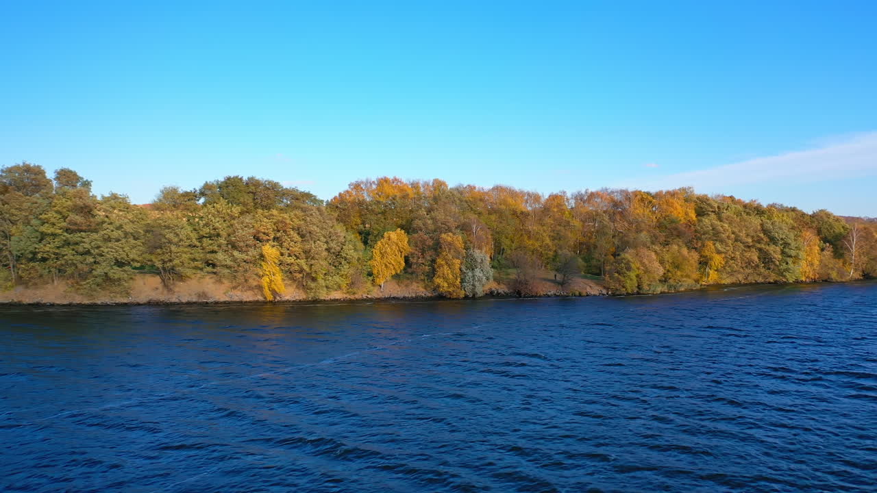 Beautiful forest near the lake under clear sky. Flight over the blue water on the background of bright autumn trees. Natural environment in fall.