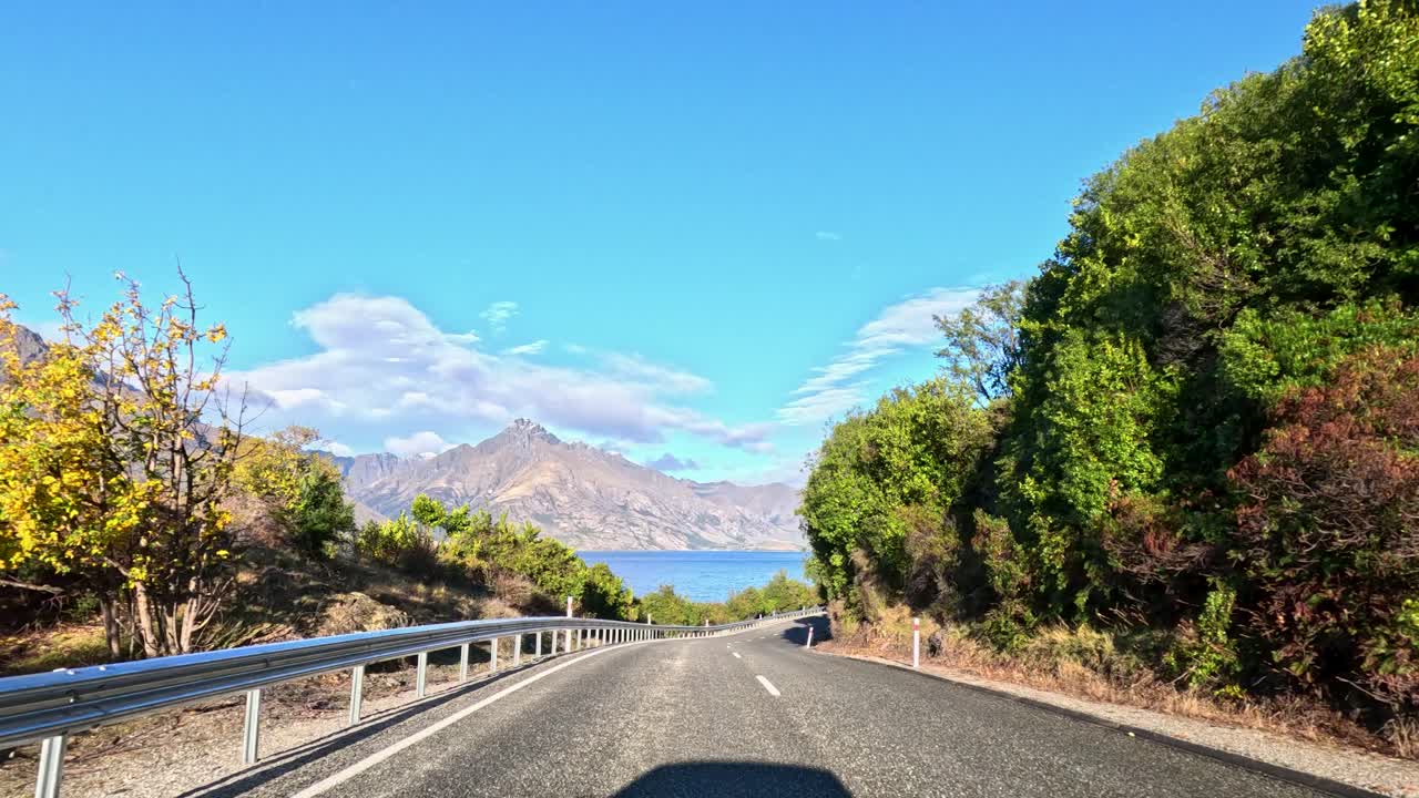 Car travels lakeside highway with mountain views, bright daylight, smooth camera motion, vibrant landscape