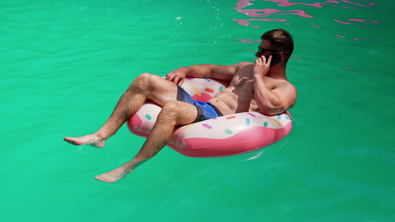 Confident relaxed Caucasian man speaks on the phone swimming in the pool. Man sits in the inflatable circle having a conversation.