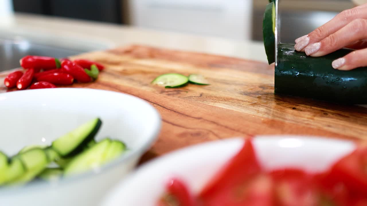 A person slices cucumber on a wooden board in a bright kitchen, surrounded by fresh vegetables