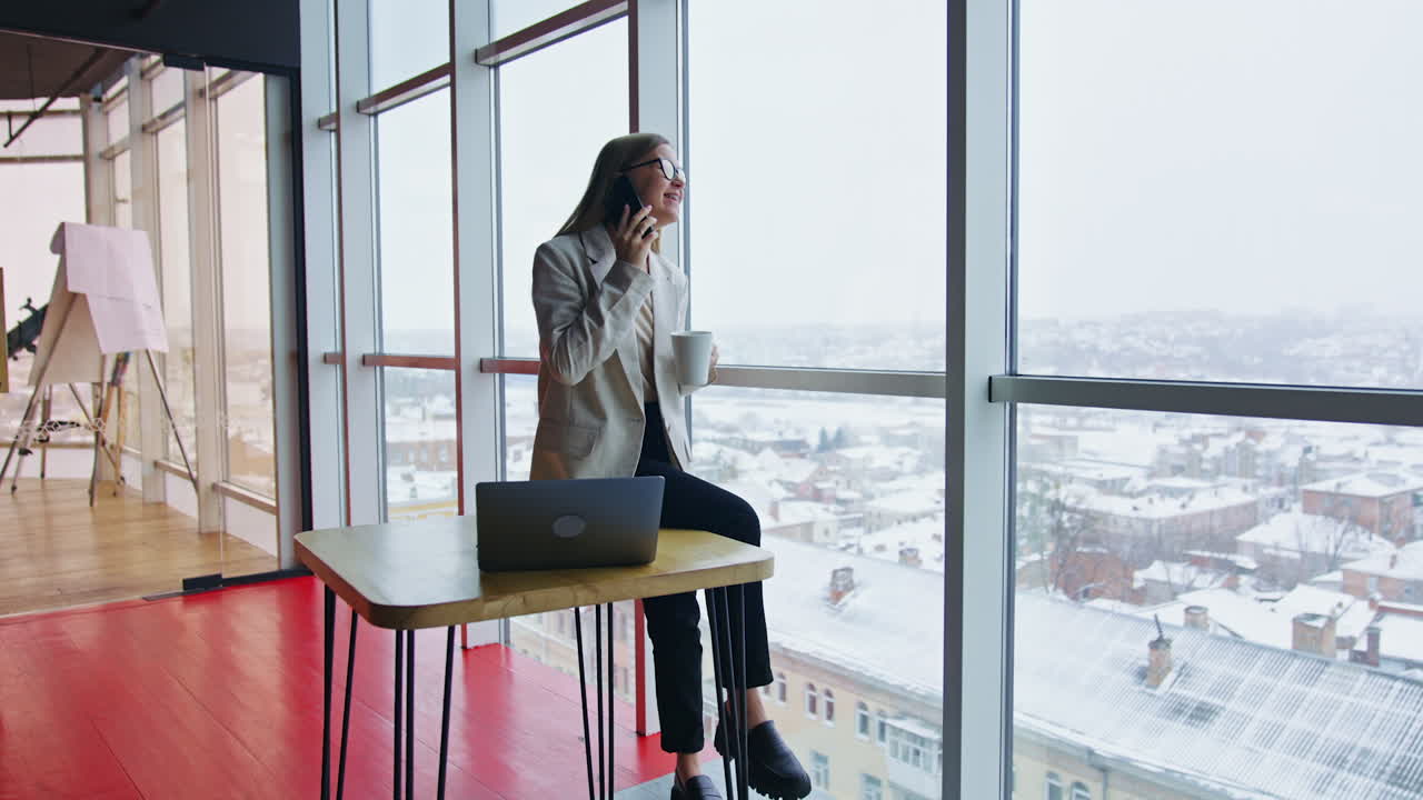 Happy female sitting on the table drinking tea and talking on the phone. Business lady having lunch break in office. Cityscape at backdrop.