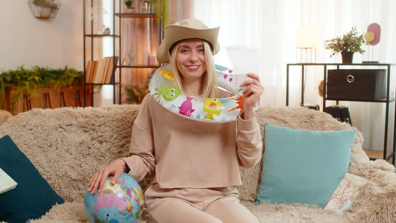 Woman sitting on sofa at home planning trip holding tickets marking route on globe with dreamy smile