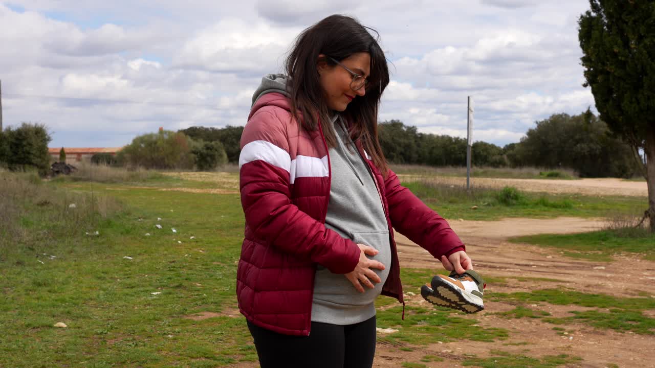 Happy pregnant woman outdoors touching belly, holding baby boots, dreaming future walks