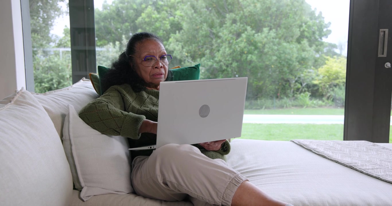 Senior African American woman sitting on sofa in green knit using laptop shifting glancing outside