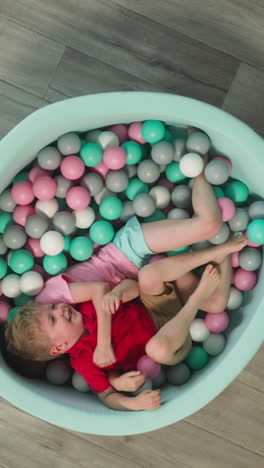 blond toddler boy has fun lying in special dry pool with elder sister. laughing children move colorful plastic balls together in cozy living room