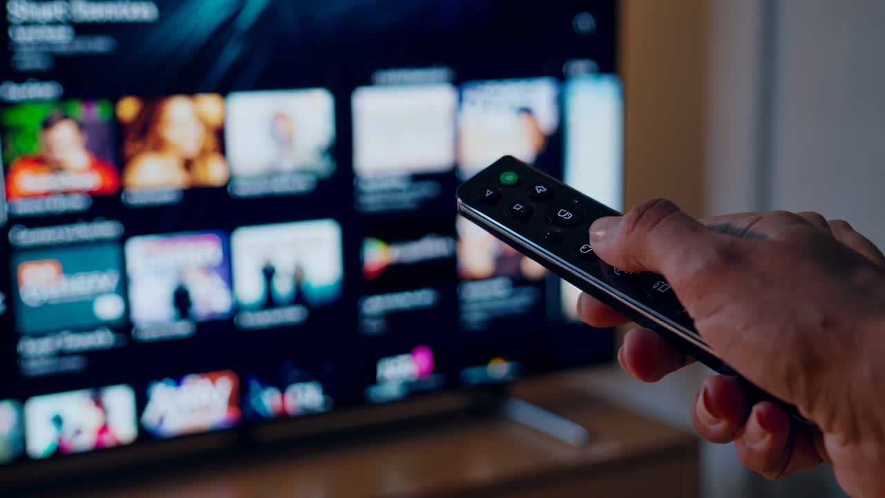 Plano Próximo de uma Mão Segurando um Controle Remoto, Apontado para uma Tela de TV Desfocada Exibindo Miniaturas de Vídeo