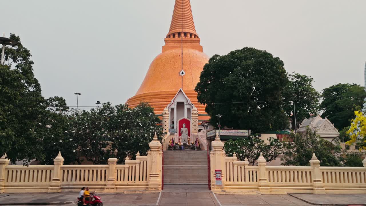 La telecamera si muove all'indietro lungo un sentiero lastricato rivelando un ampio ingresso che conduce verso una gigantesca stupa arancione, mentre la luce mattutina velata illumina gli alberi verdi che circondano il sacro sito buddista in Thailandia