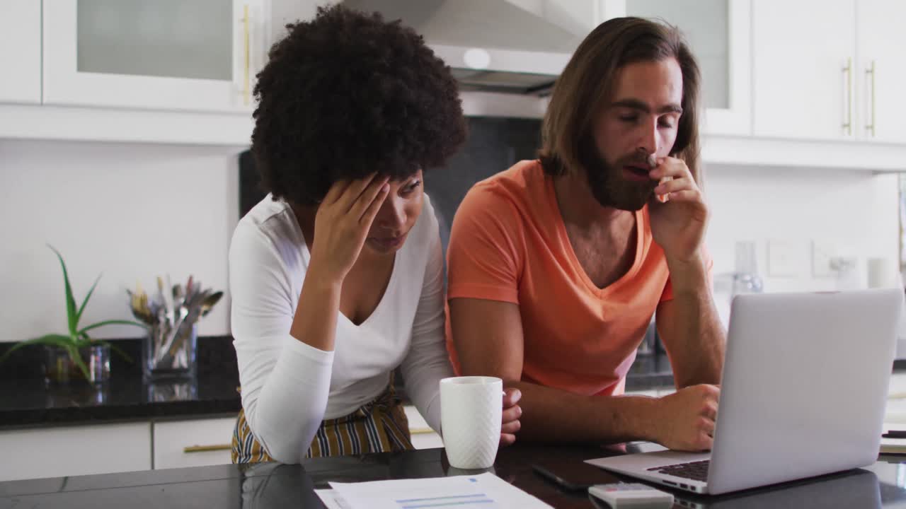 Stressed mixed race couple using laptop and calculating finances in the kitchen at home