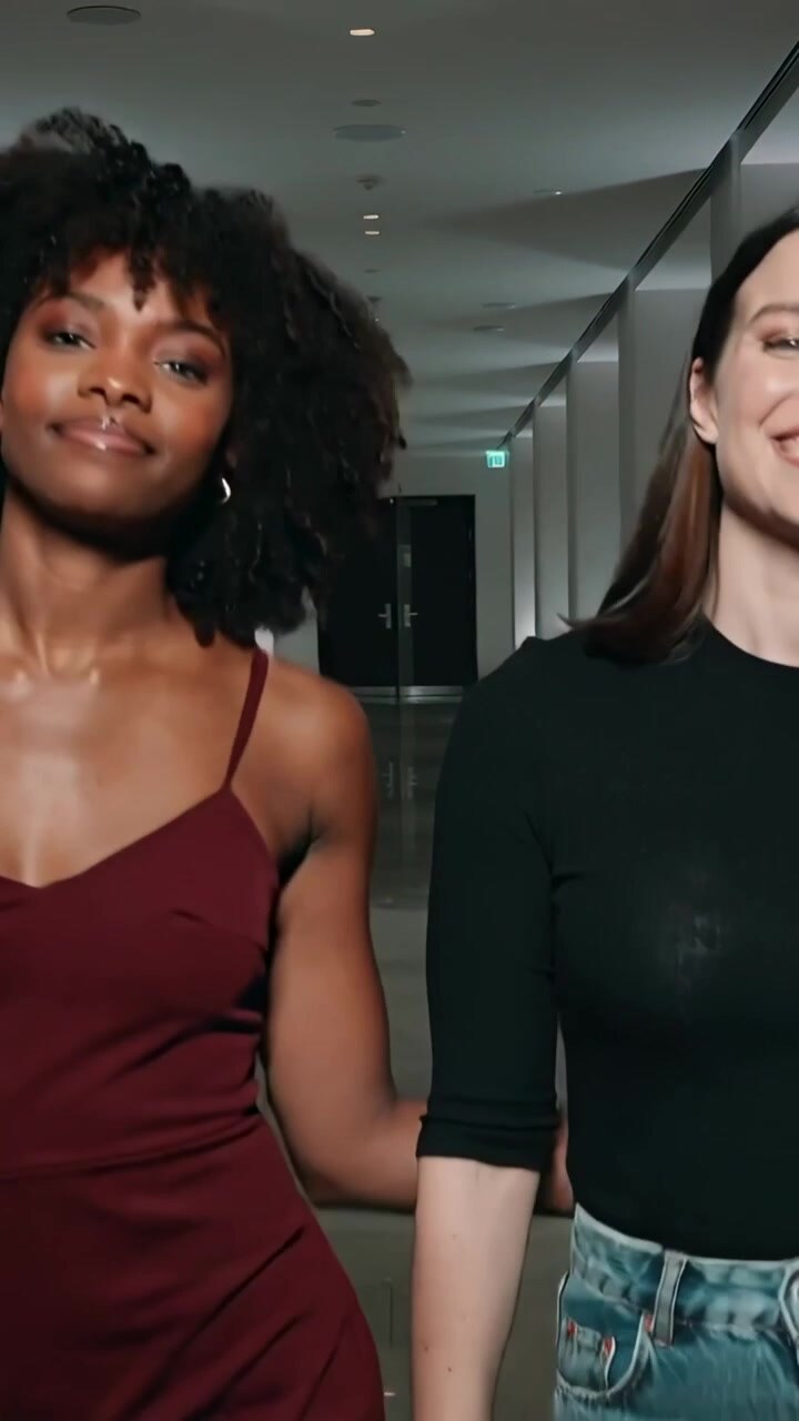 Video verticale: Due donne a braccetto che camminano in un corridoio, salutando la telecamera, una in vestito bordeaux e l'altra in jeans