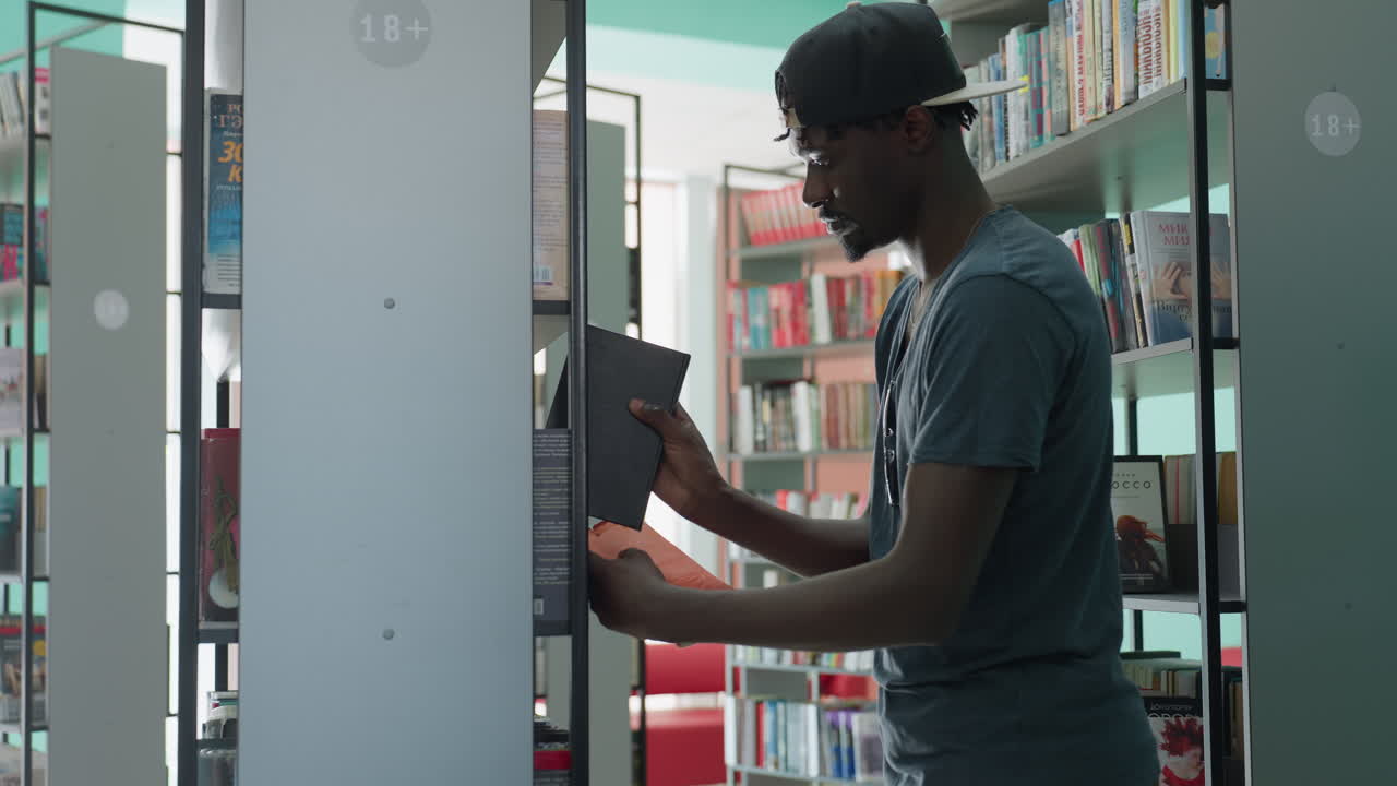 Young man in casual t-shirt and cap standing in bright library, extending arm to return book to shelf, surrounded by tall bookcases filled with colorful titles