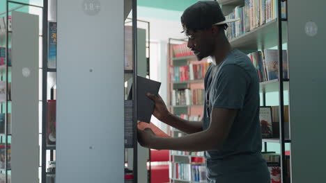 Young man in casual t-shirt and cap standing in bright library, extending arm to return book to shelf, surrounded by tall bookcases filled with colorful titles