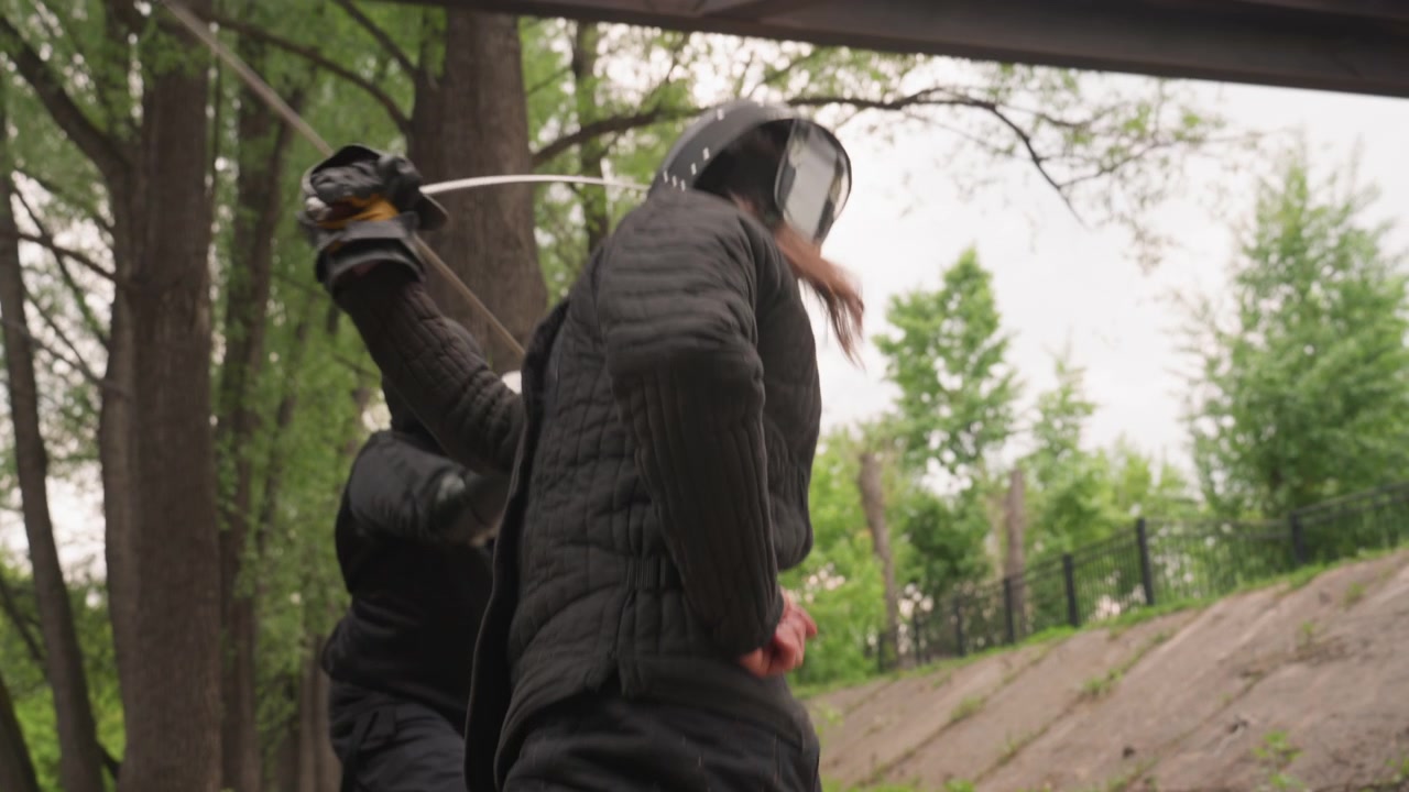 Two people in black padded armor engage in sword fight outdoors with one person wielding sword while other prepares to defend. scene takes place on uneven ground surrounded by trees