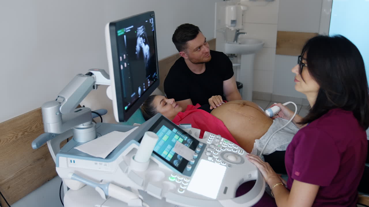 Female dark-haired doctor sits in front of ultrasound machine. Medic performs check up for a pregnant woman. Man sits beside his wife.